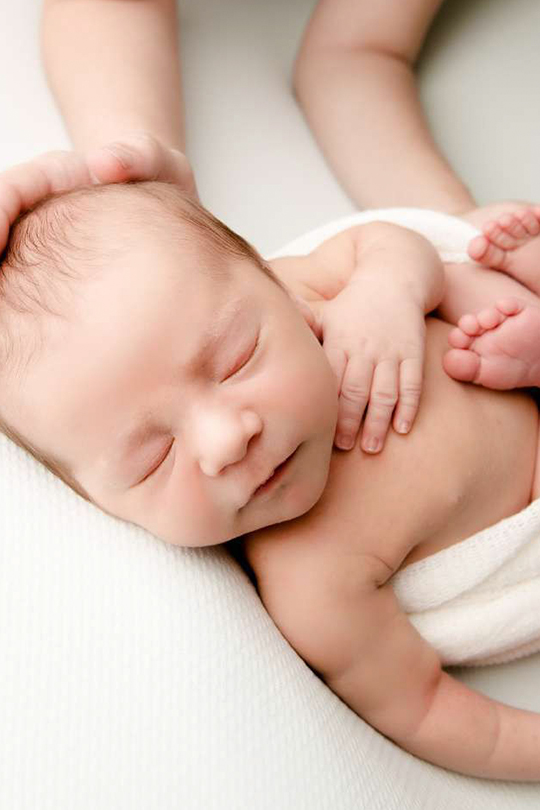 Un recién nacido descansa plácidamente sobre una superficie blanca con los ojos cerrados. Una manita descansa sobre su pecho, mientras una mano de adulto acuna suavemente la cabeza del bebé. El bebé está envuelto en una suave manta blanca. Un recién nacido descansa plácidamente sobre una superficie blanca con los ojos cerrados. Una manita descansa sobre su pecho, mientras una mano de adulto acuna suavemente la cabeza del bebé. El bebé está envuelto en una suave manta blanca.