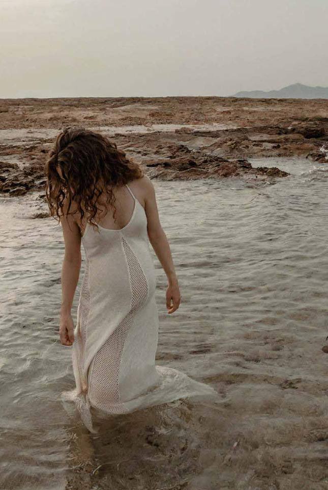 Una persona de pelo rizado con un vestido blanco transparente está de pie en aguas poco profundas, en una orilla rocosa, mirando hacia abajo con el rostro parcialmente oculto. El cielo está nublado y se ven montañas a lo lejos. Una persona de pelo rizado con un vestido blanco transparente está de pie en aguas poco profundas, en una orilla rocosa, mirando hacia abajo con el rostro parcialmente oculto. El cielo está nublado y se ven montañas a lo lejos.